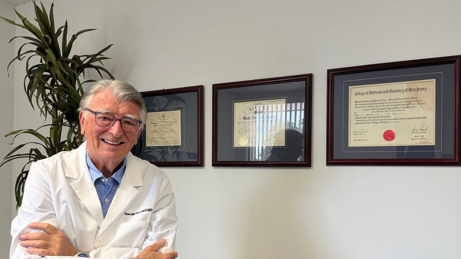 Dr. Carroll, arms folded, standing in front of his framed certificates at the Maitland office