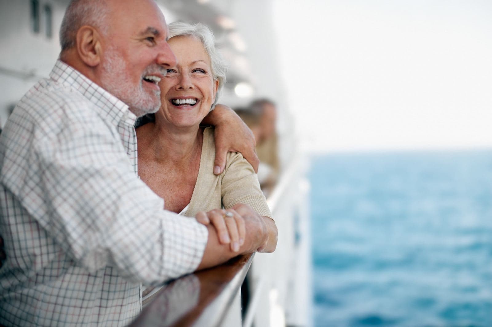 An older couple leaning on a ship rail, laughing, with the sea behind them